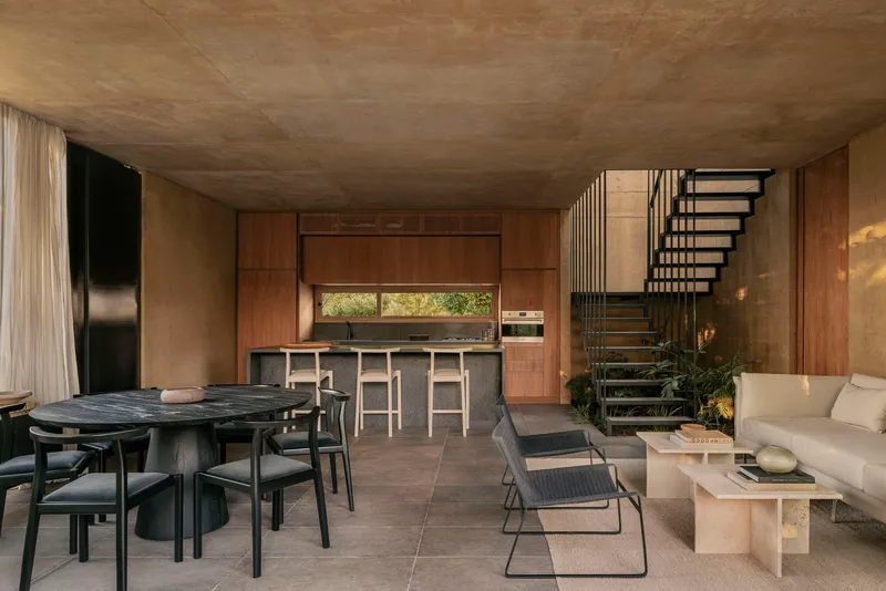 Open-plan living space with exposed concrete ceiling, timber kitchen and metal staircase beside dining area