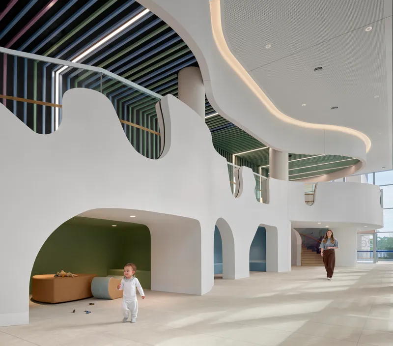 Atrium with curved white walls, arched openings, and rainbow-striped ceiling baffles with child running below