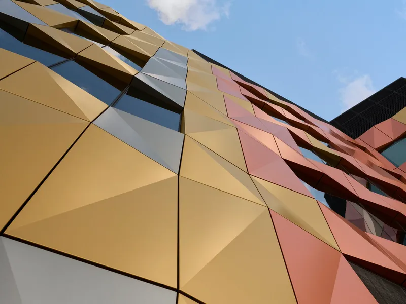 Upward view of the geometric metal panel facade in gold, pink, and copper tones against a blue sky