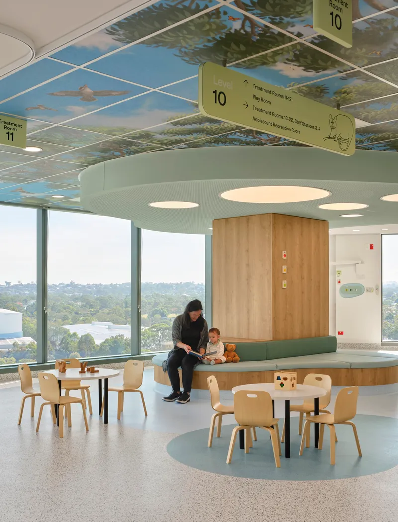 Seating area with timber volume, plywood furniture, and ceiling mural of trees with hanging wayfinding signs