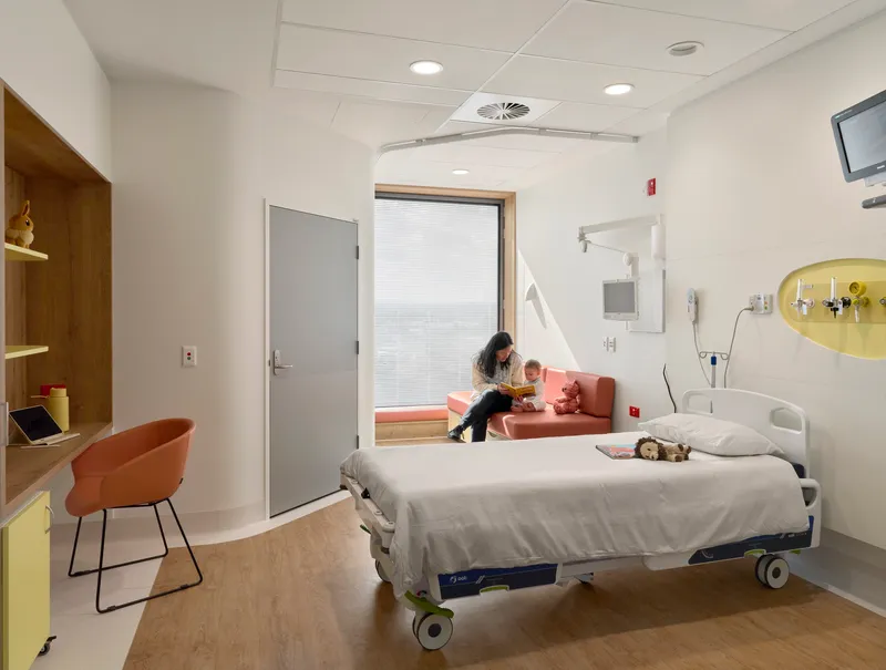 Patient room with hospital bed, window seat, and wooden desk under a suspended ceiling with recessed lighting