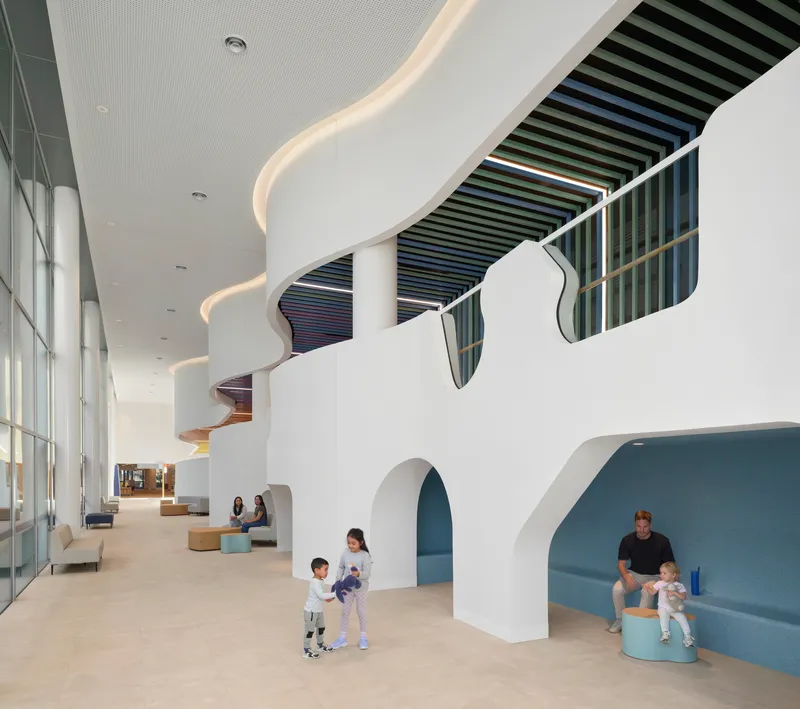 Double-height atrium with organic curving openings, colorful baffled ceiling and families seated in arched alcoves below
