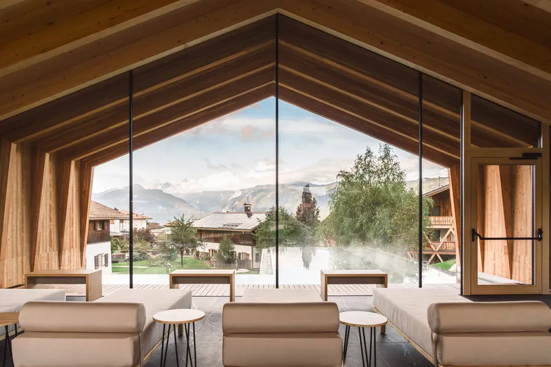 Lounge space beneath timber beam ceiling framing a view through glazing to mist-covered pool terrace