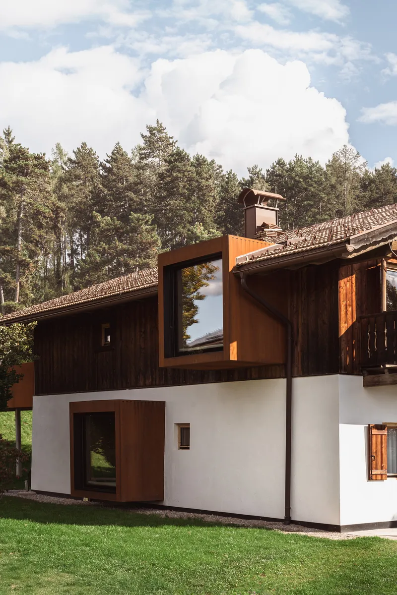 White stucco base with weathered steel bay windows projecting from dark timber upper floor under cloudy sky
