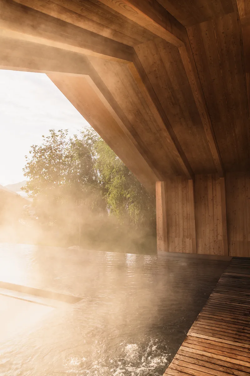 Cantilevered timber roof over steaming pool with angled wood beams in morning mist