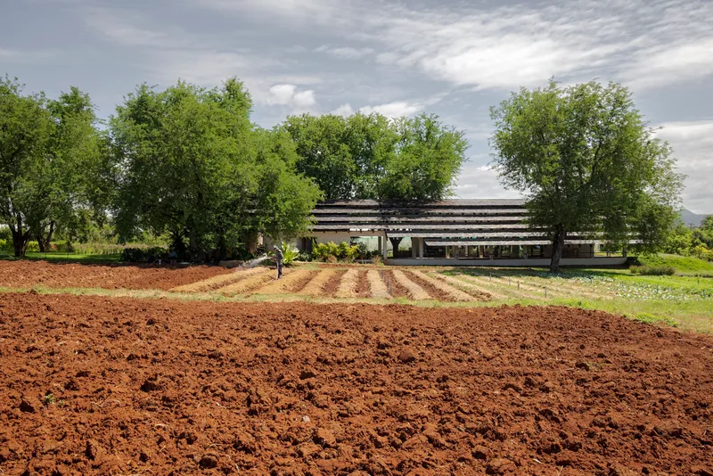 Longitudinal elevation with stacked timber eaves seen across freshly tilled red earth and a figure
