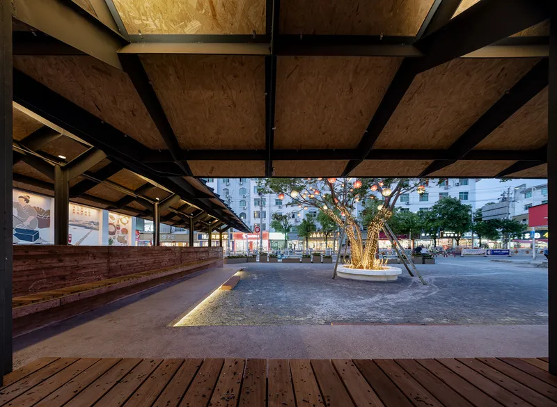 Covered timber deck with plywood ceiling panels and black steel beams overlooking a plaza at twilight