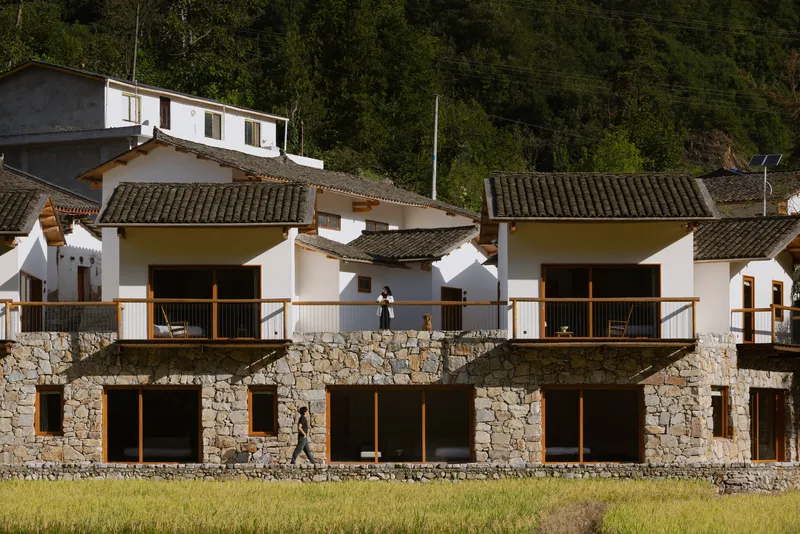 Two-story facade with stone base and white upper walls fronting a rice field in bright sunlight
