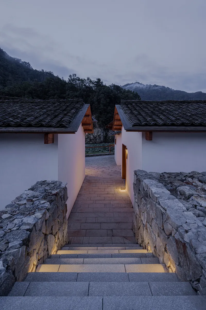 Lit stairway descending between stone retaining walls toward white pavilions under darkening mountain sky
