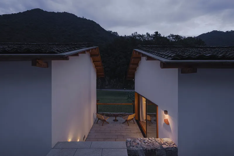 Courtyard passage at dusk with uplights along stone walls and two chairs beside glazed opening