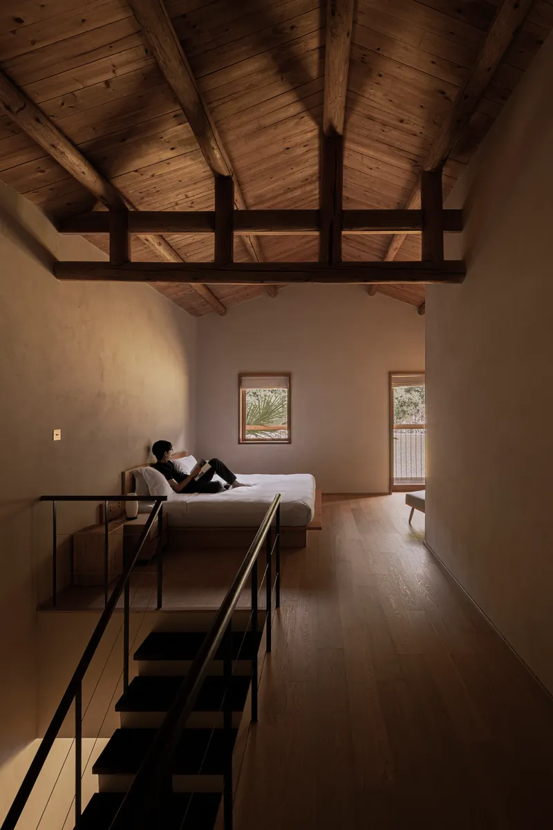 Double-height bedroom with exposed timber roof trusses and a person reading on the daybed