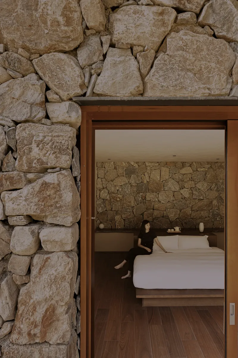 Open doorway through dry-stacked stone wall revealing bedroom with matching stone headboard wall