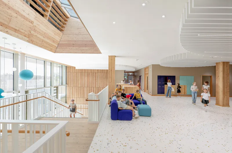 Double-height gathering space with angular timber skylight, timber slat walls and children on soft seating