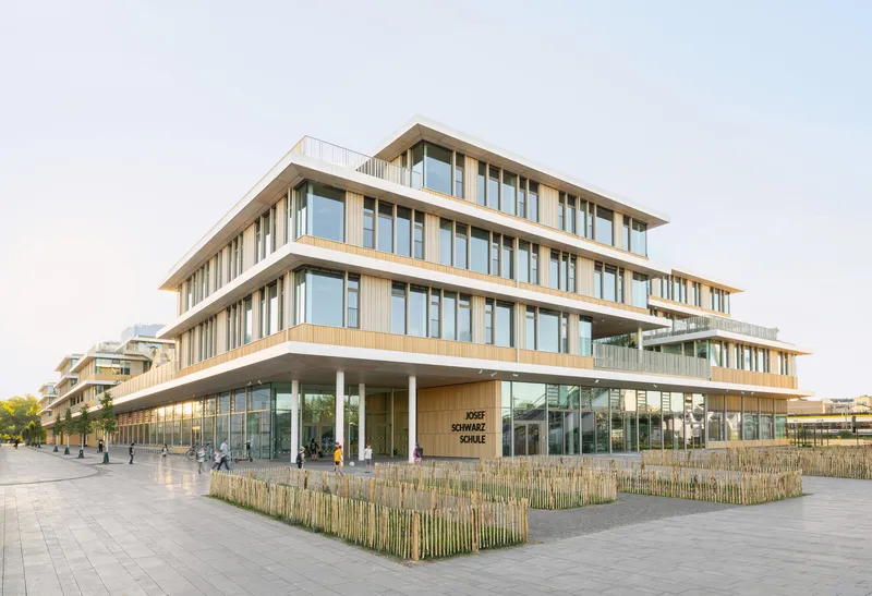 Corner view of the timber and glass facade with stacked floor plates and planted beds at the plaza
