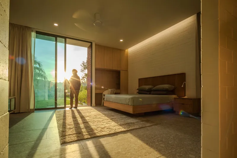 Bedroom interior with wood-paneled walls and full-height glazing framing two figures in golden sunset light