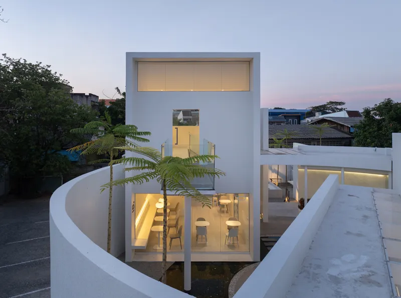 Multi-level white facade with stacked glass openings and curved perimeter wall beside a young tree at dusk