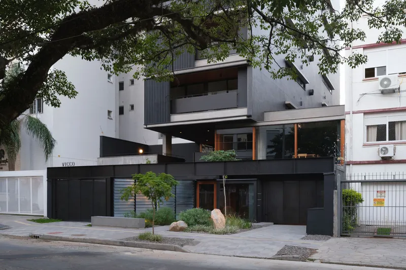 Street facade with black horizontal volumes and dark corrugated metal tower framed by overhanging tree branches