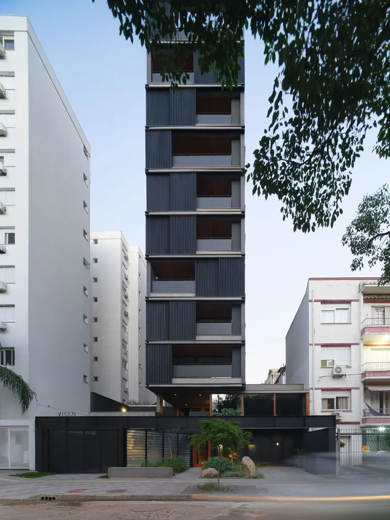 Slender tower facade with offset balconies and vertical corrugated cladding photographed at dusk beneath tree foliage