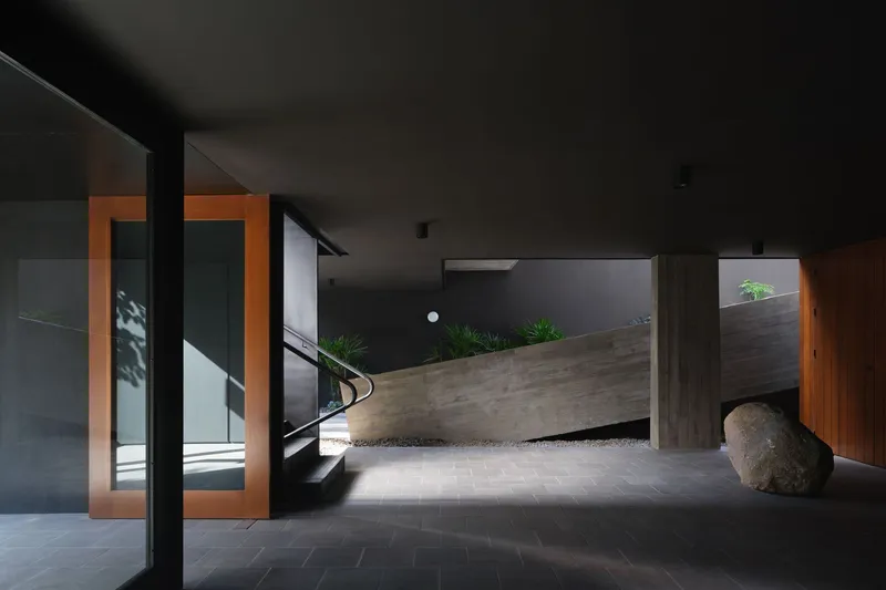 Entry foyer with angled concrete wall, timber-framed doorways casting orange light, and a large boulder