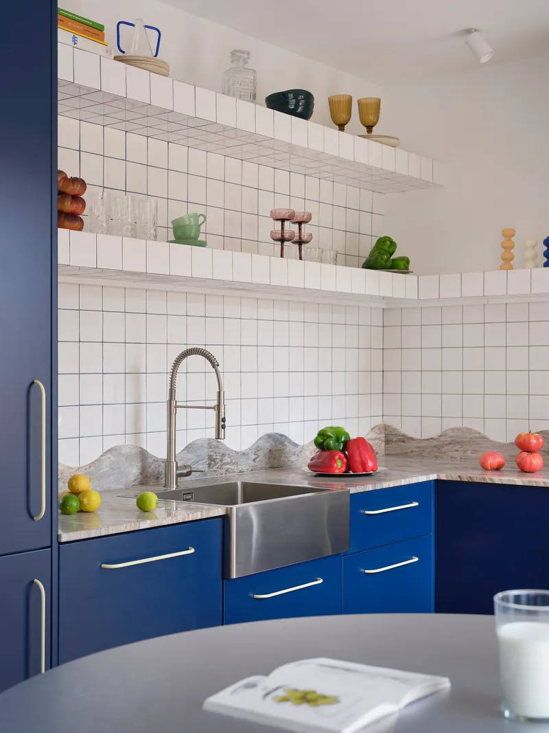 Corner kitchen with blue cabinets, white square tile backsplash, and open shelves displaying glassware and plants