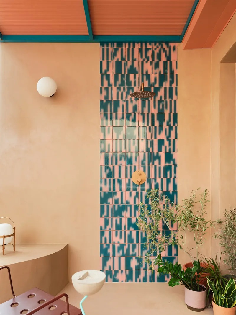 Bathroom shower wall with pink and teal mosaic tile beneath peach ceiling and potted plants at floor