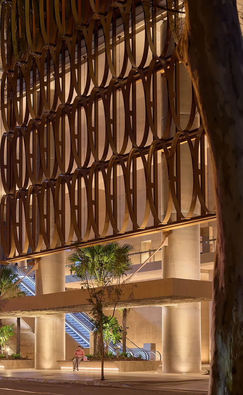 Multi-story atrium facade with woven timber screen pattern and palm trees at ground level at dusk