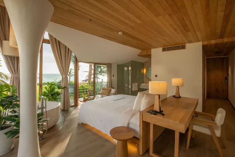 Bedroom with freestanding bathtub and timber-framed glazing opening to balcony overlooking ocean horizon at dusk