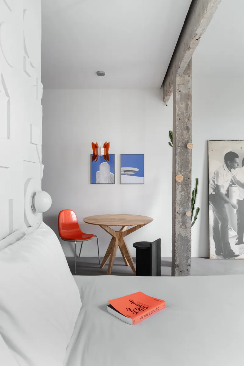 Sitting nook with round timber table, red chair, and artwork against a white wall beside concrete column