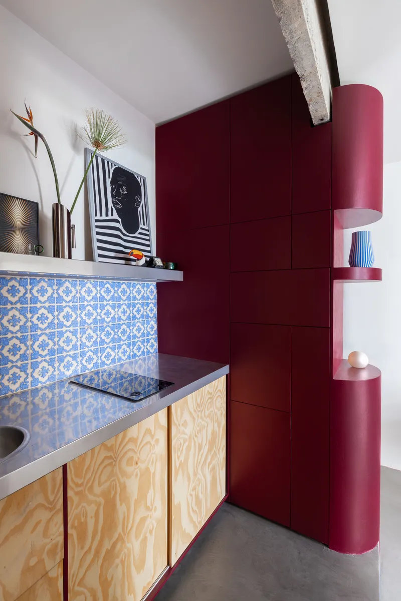 Kitchenette with blue patterned tile backsplash, plywood cabinets, and burgundy cylindrical storage column