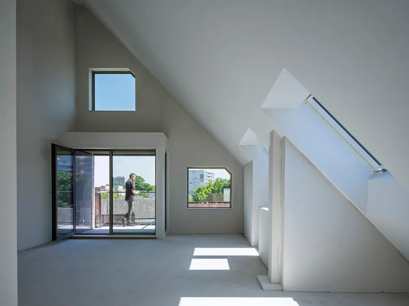 Interior attic space with sloped ceilings, multiple windows, and a figure on the balcony in afternoon light