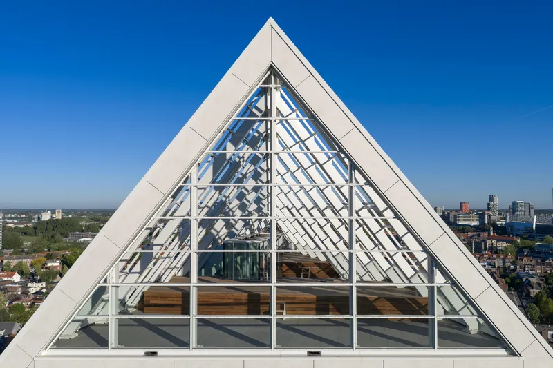 Triangular glass gable with exposed white structural frame against a clear blue sky and urban horizon