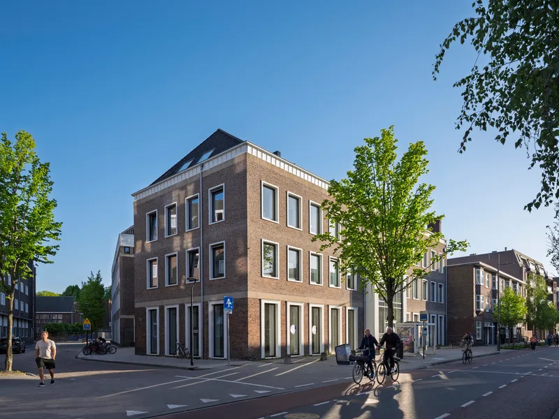 Street corner view of brick volume with white trim and planted trees with cyclists passing
