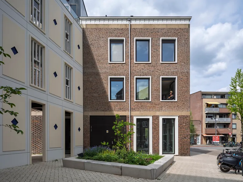 Brown brick facade with symmetrical white-framed windows and planted terrace beside pale neighboring facade