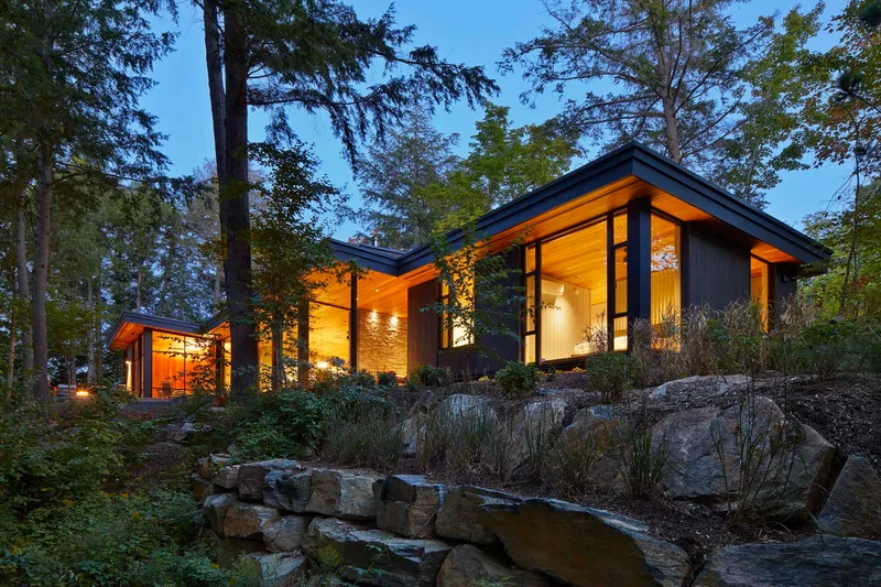 Illuminated residence with sloping roofline among boulders and native planting at twilight