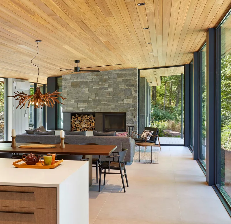Open living space with stone fireplace, timber ceiling, and floor-to-ceiling windows framing woodland views