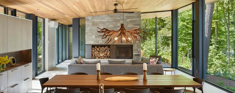 Open living and dining space with timber ceiling and stone fireplace framed by forest views
