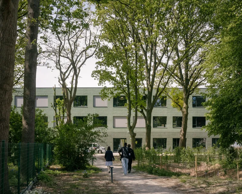 View through mature trees of the green tiled facade with two students walking on a gravel path