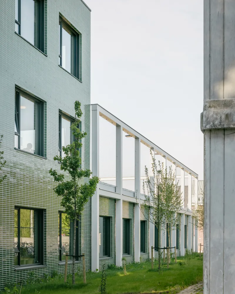 Courtyard passage between green tile facade and white colonnade with young trees at dusk