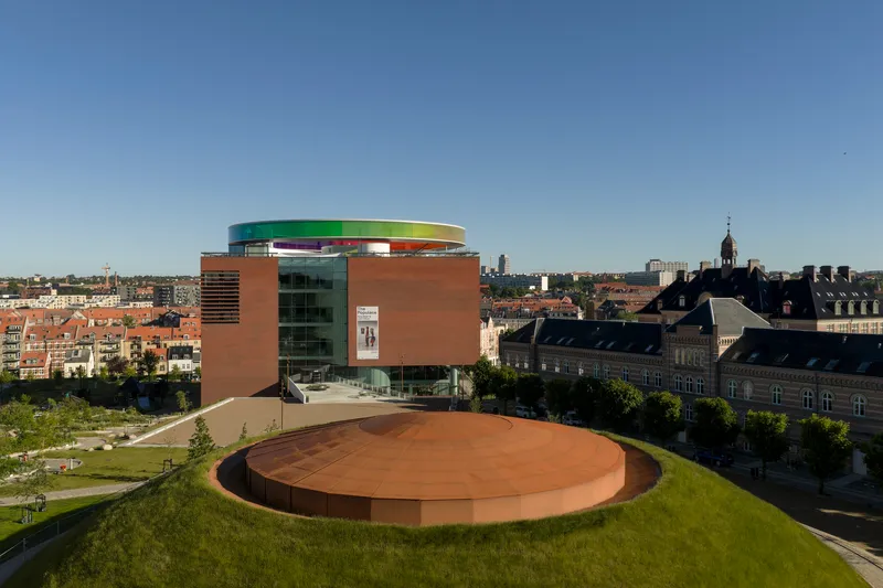 Cylindrical building with terracotta panels and rainbow rooftop overlooking the surrounding urban district