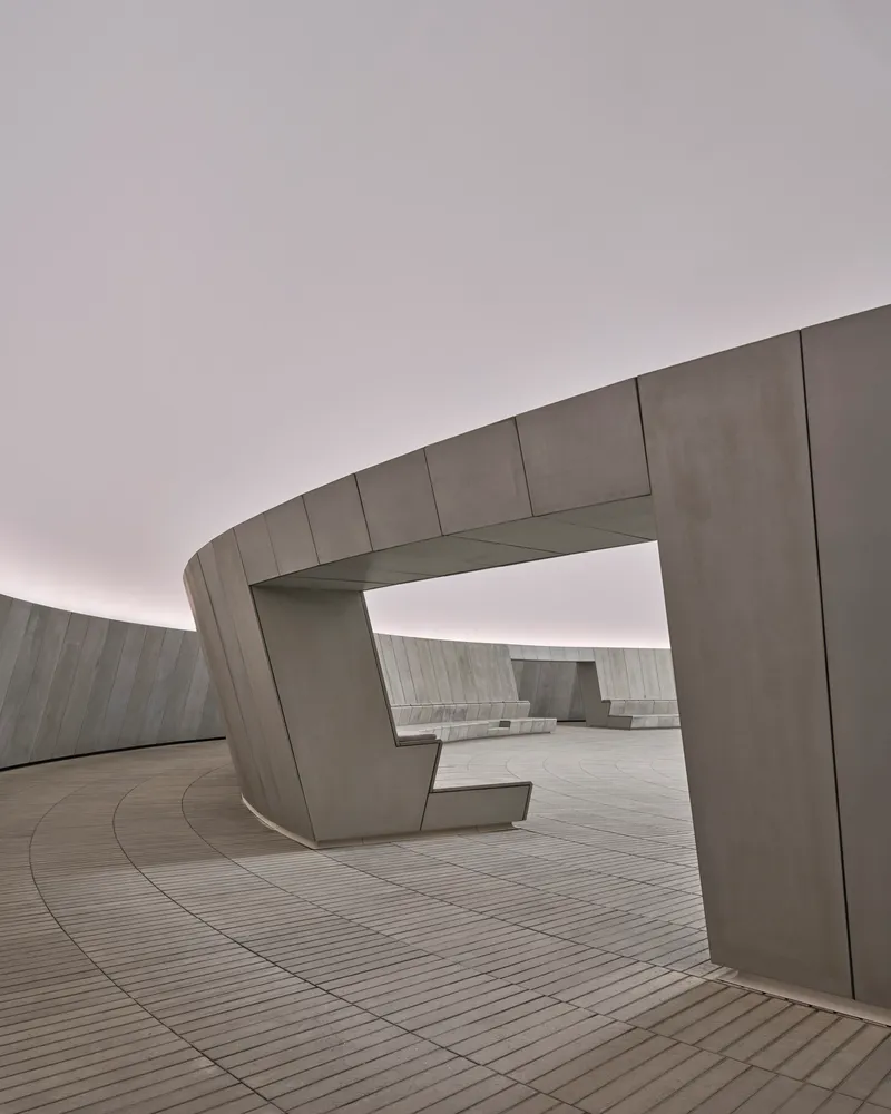 Precast concrete seating elements with angular forms arranged along the curved perimeter wall under cloudy skies