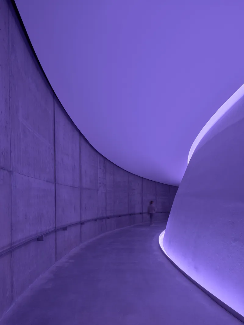 Curved concrete corridor with purple light washing the ceiling and a solitary figure walking