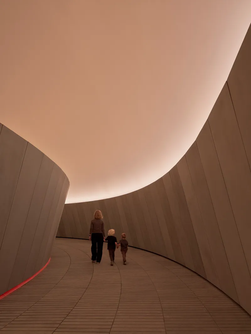 Ramped passageway with concrete walls and warm overhead light as visitors ascend together