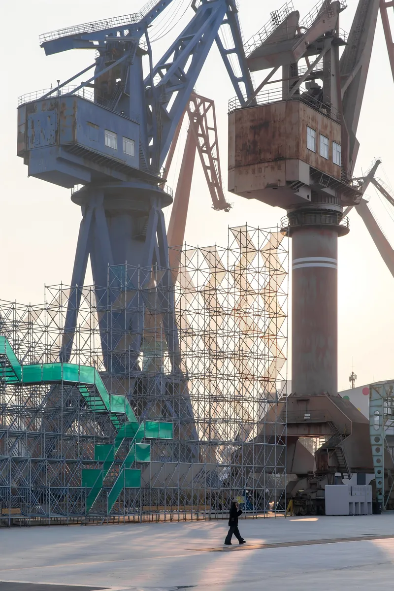 Ground view of the scaffolding structure with existing harbor cranes and a pedestrian at dusk
