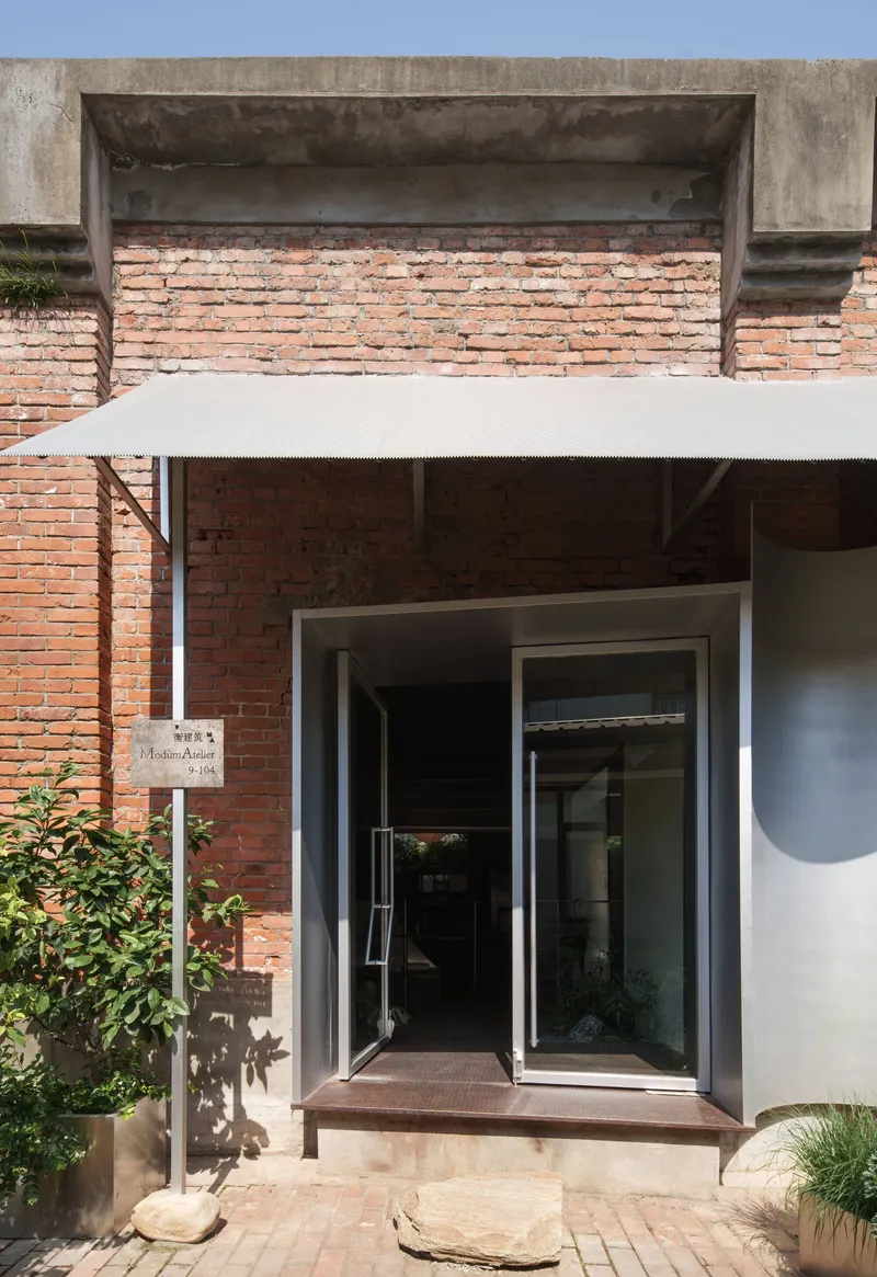 Recessed entrance with exposed brick wall, concrete lintel and glazed pivot door under white canopy