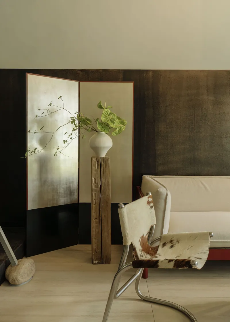 Corner vignette showing folding screen, timber pedestal with vase and cowhide chair in soft light