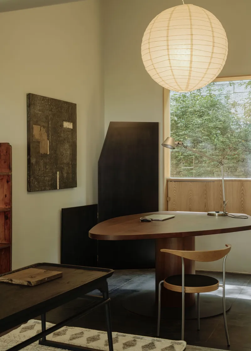 Workspace with oval table, paper lantern and timber casement window framing courtyard vegetation