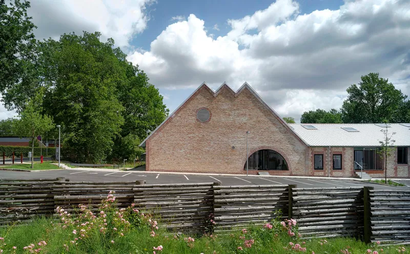 Triple-gabled brick facade with arched entrance behind a low woven timber fence and planted bed