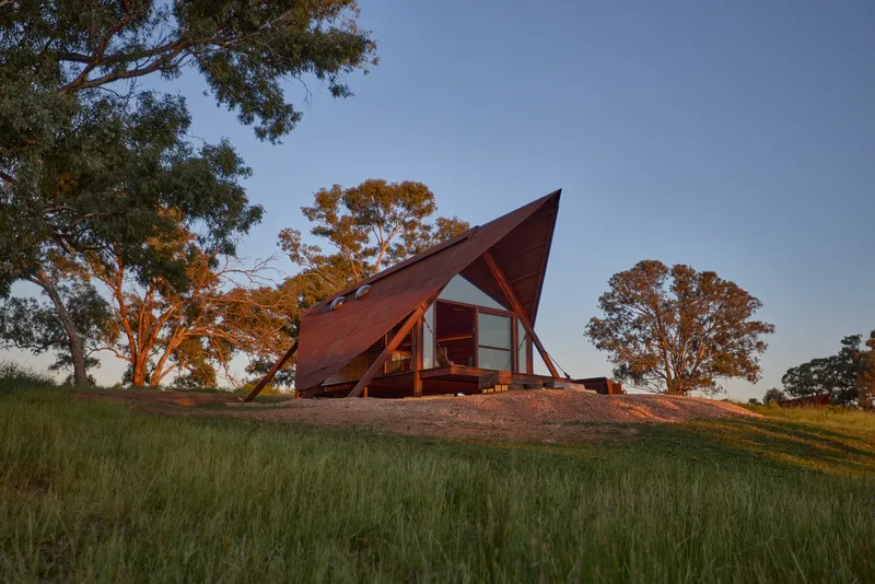 A-frame cabin with weathered steel cladding on a grassy hillside at dusk beneath eucalyptus trees
