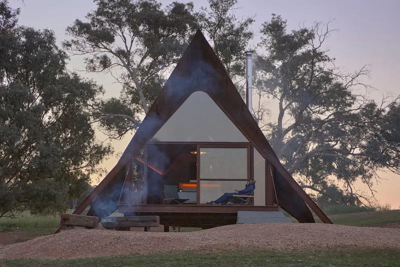Triangular timber cabin with glazed facade and smoking chimney among eucalyptus trees at twilight