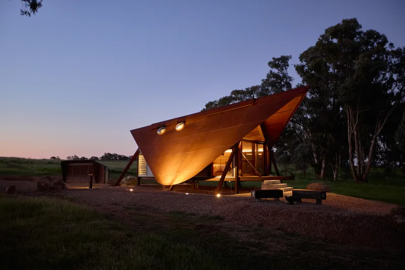 Tilted angular timber volume with circular windows glowing at dusk beside a grove of eucalyptus trees